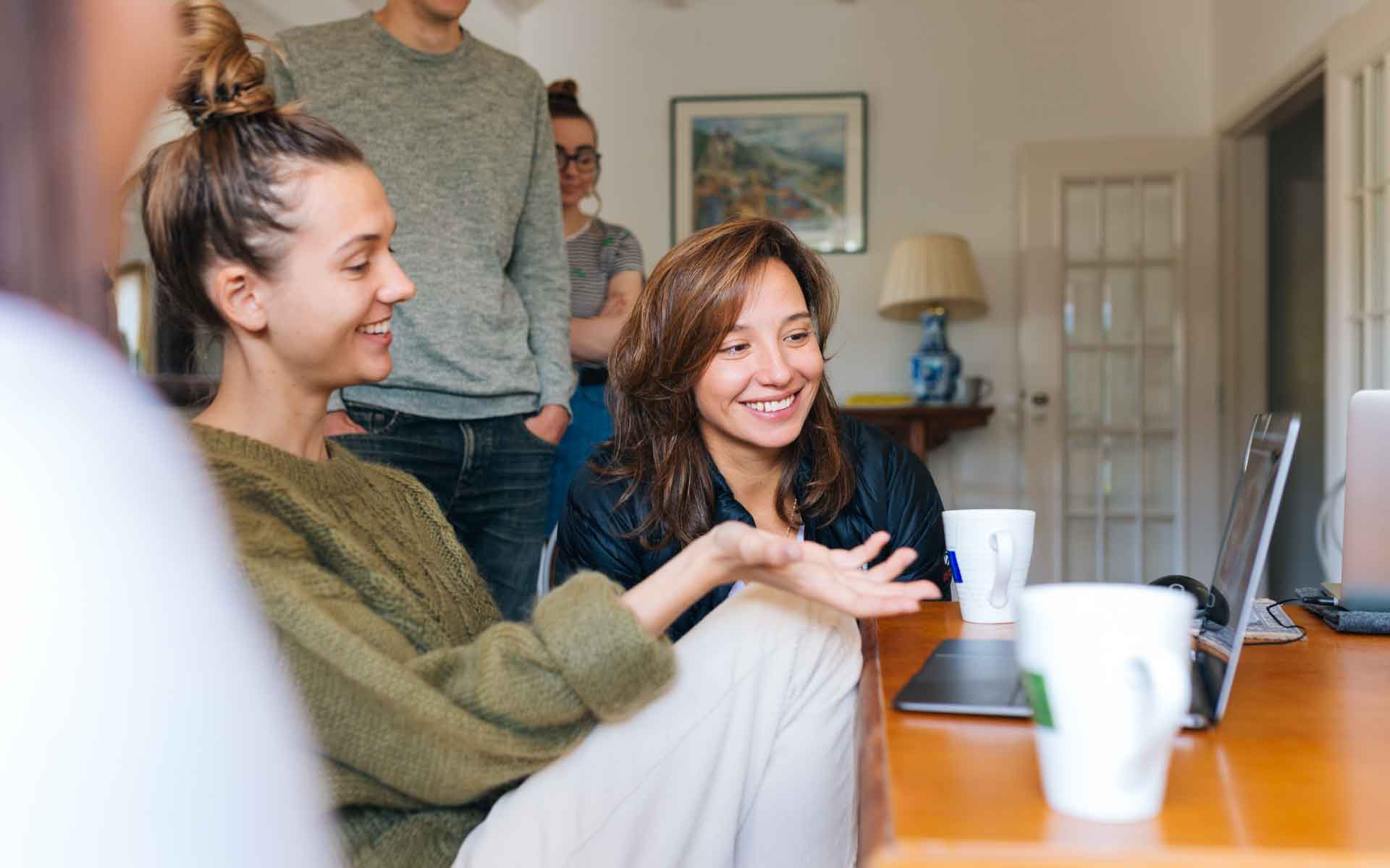 Group of young adults engaging in discussion around a laptop, with coffee cups on a table, reflecting collaboration and decision-making related to home selling and finances.