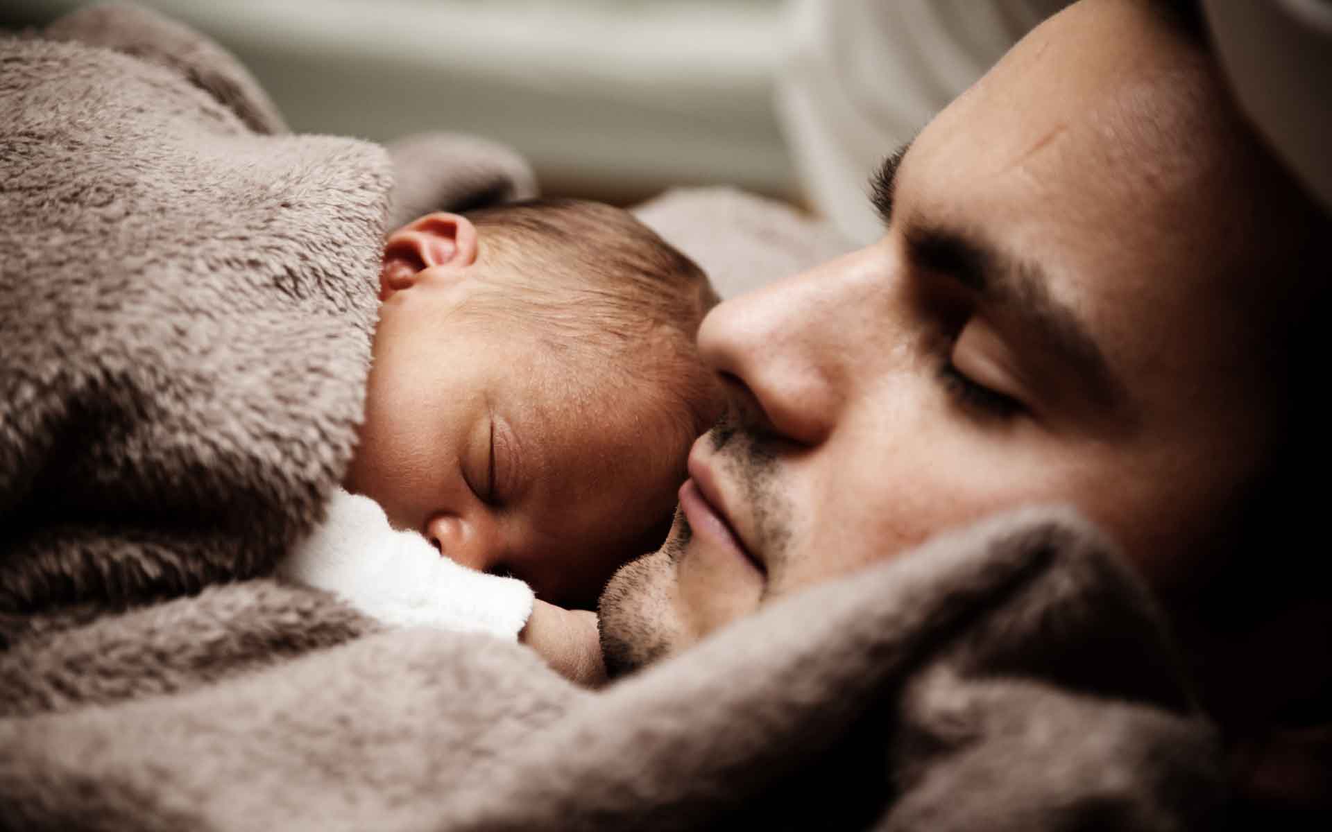 Father and newborn baby peacefully sleeping together, wrapped in soft blankets, illustrating the comfort of family life.