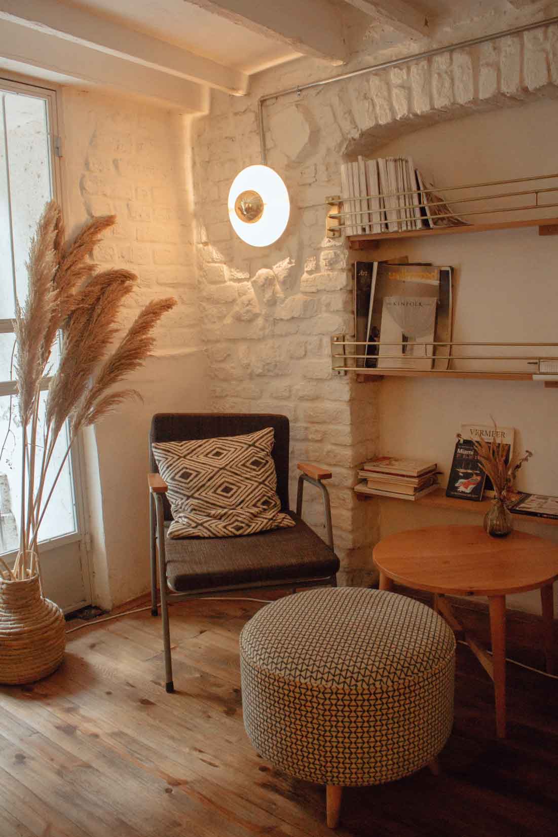 Cozy reading nook featuring a chair with a patterned pillow, a round ottoman, a decorative vase, and a shelf with magazines, set against a light brick wall.