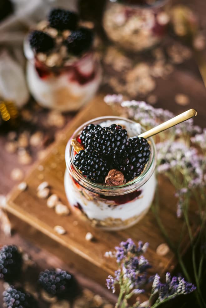 Blackberry parfait in a glass jar with granola and yogurt, surrounded by fresh blackberries and lavender flowers, showcasing a healthy dessert option.