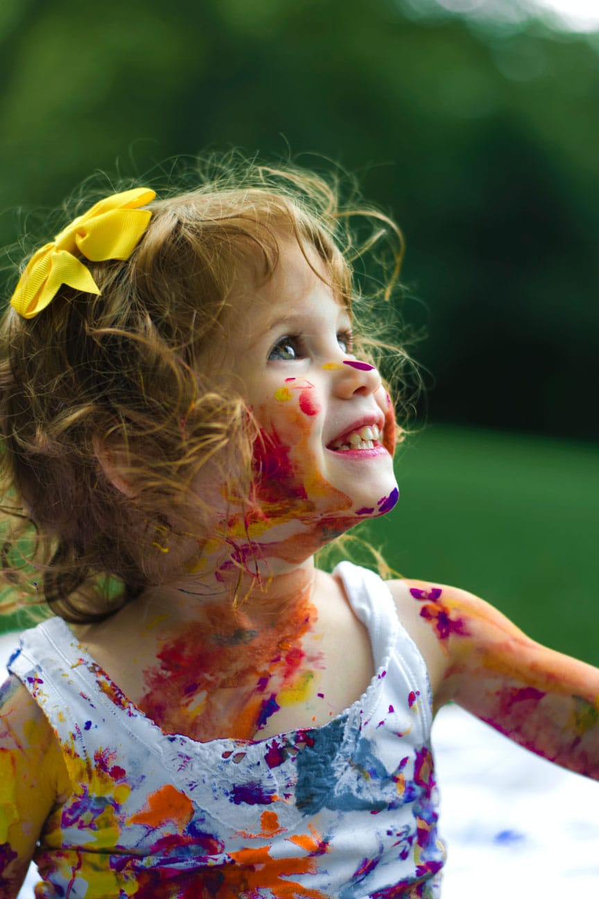 Girl with curly hair and yellow bow, smiling joyfully, covered in colorful paint, outdoors.