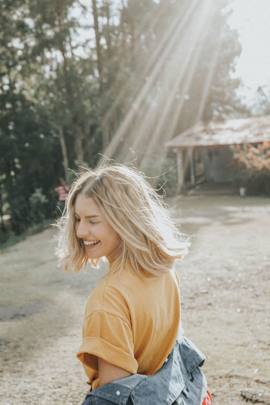 Smiling young woman in a yellow shirt, enjoying the outdoors with sunlight streaming through trees, representing a carefree lifestyle relevant to home buying and rental property insurance.