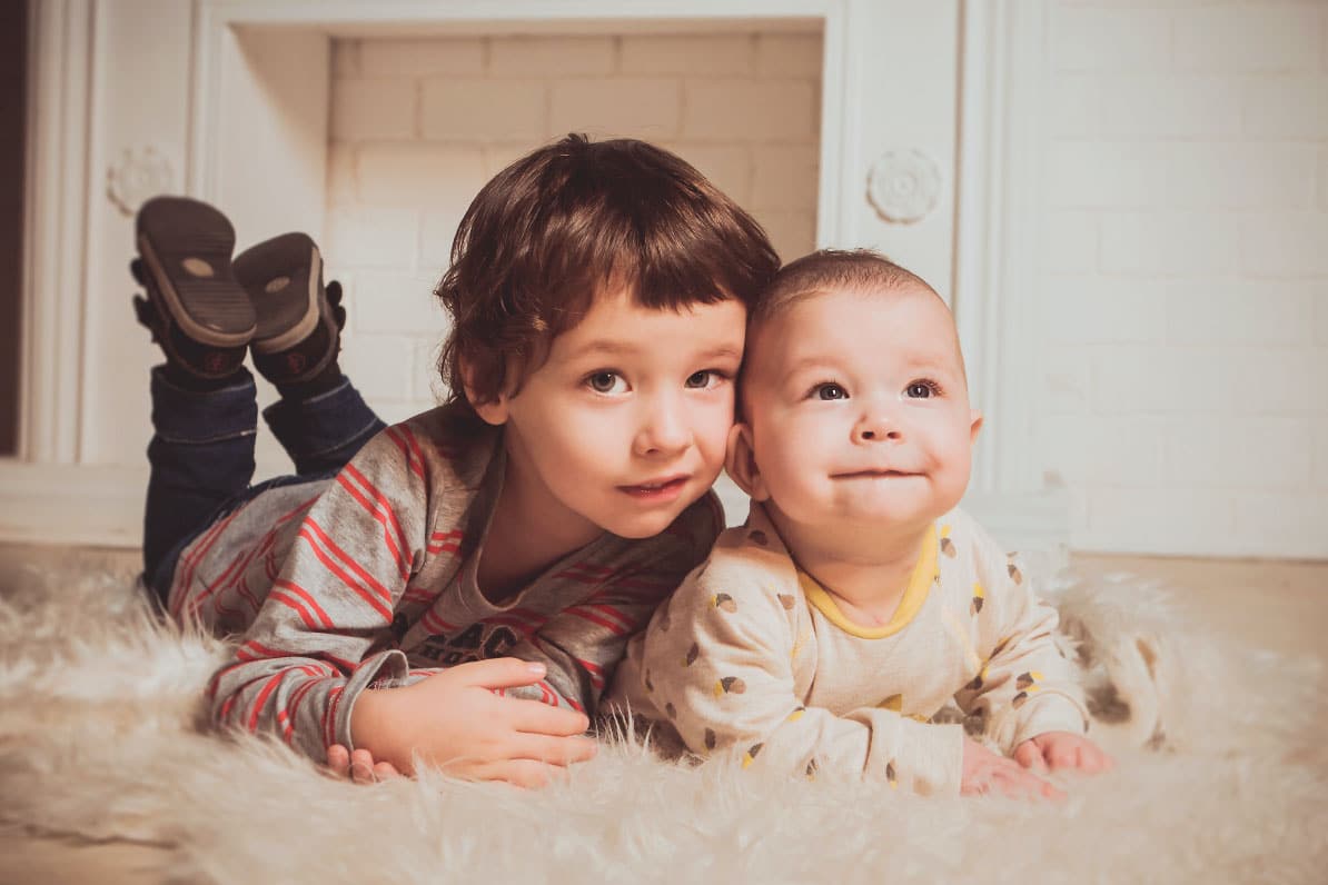 Two children, a young boy and a baby, lying on a soft rug in a cozy indoor setting, smiling and enjoying each other's company.