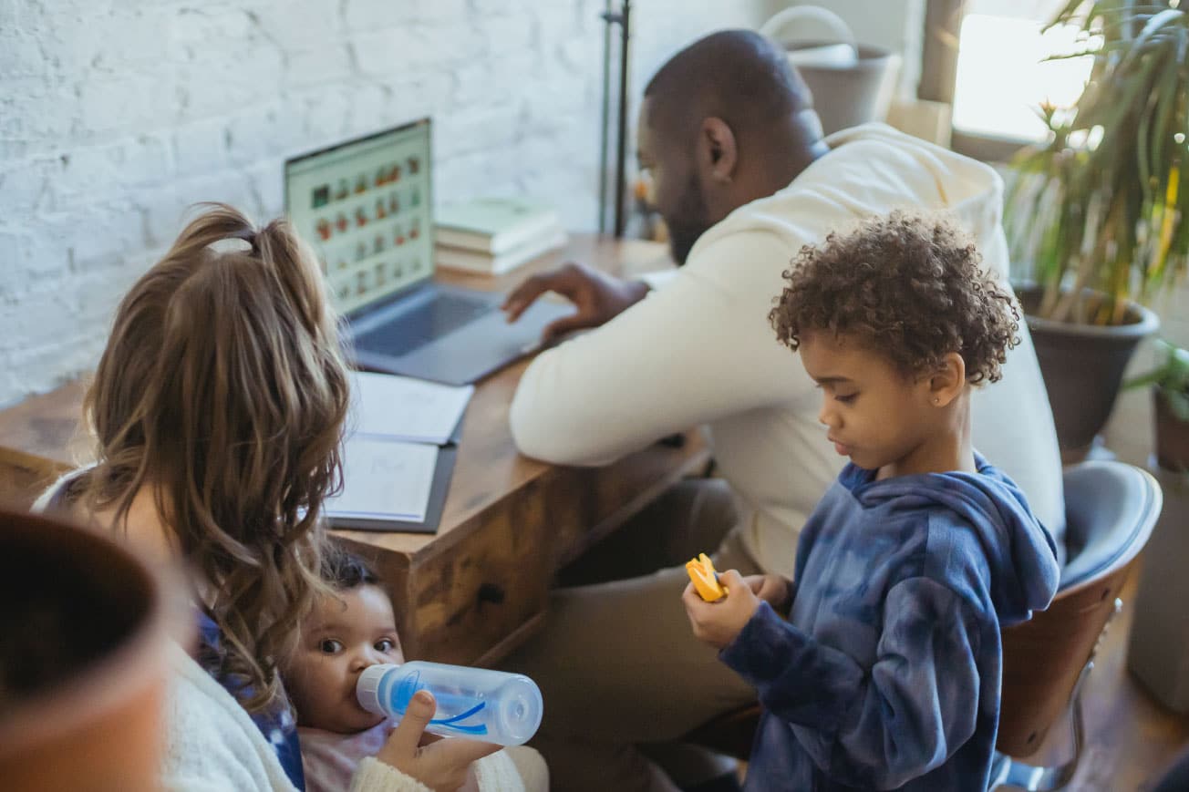Family engaging in home buying research at a wooden table, with a mother feeding a baby, a father using a laptop, and a child holding a snack, emphasizing the home buying process and family involvement.
