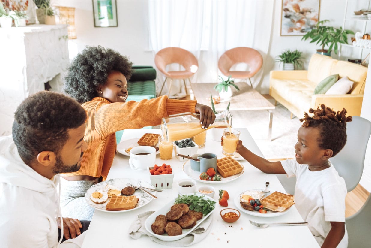 Family enjoying breakfast together, featuring waffles, fruits, and drinks, as a woman pours orange juice for a child in a bright, modern dining space.
