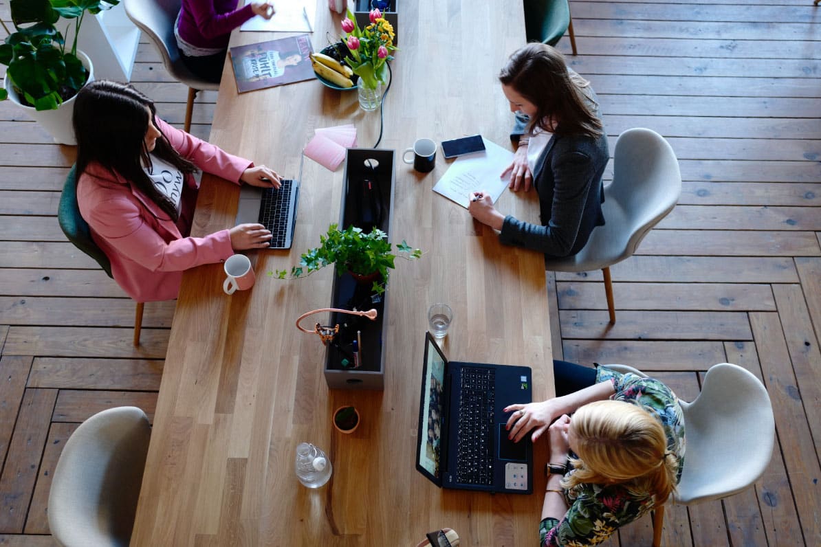 Group of women collaborating at a wooden table, using laptops and taking notes, with plants and coffee cups, in a workspace setting related to real estate investment discussions.