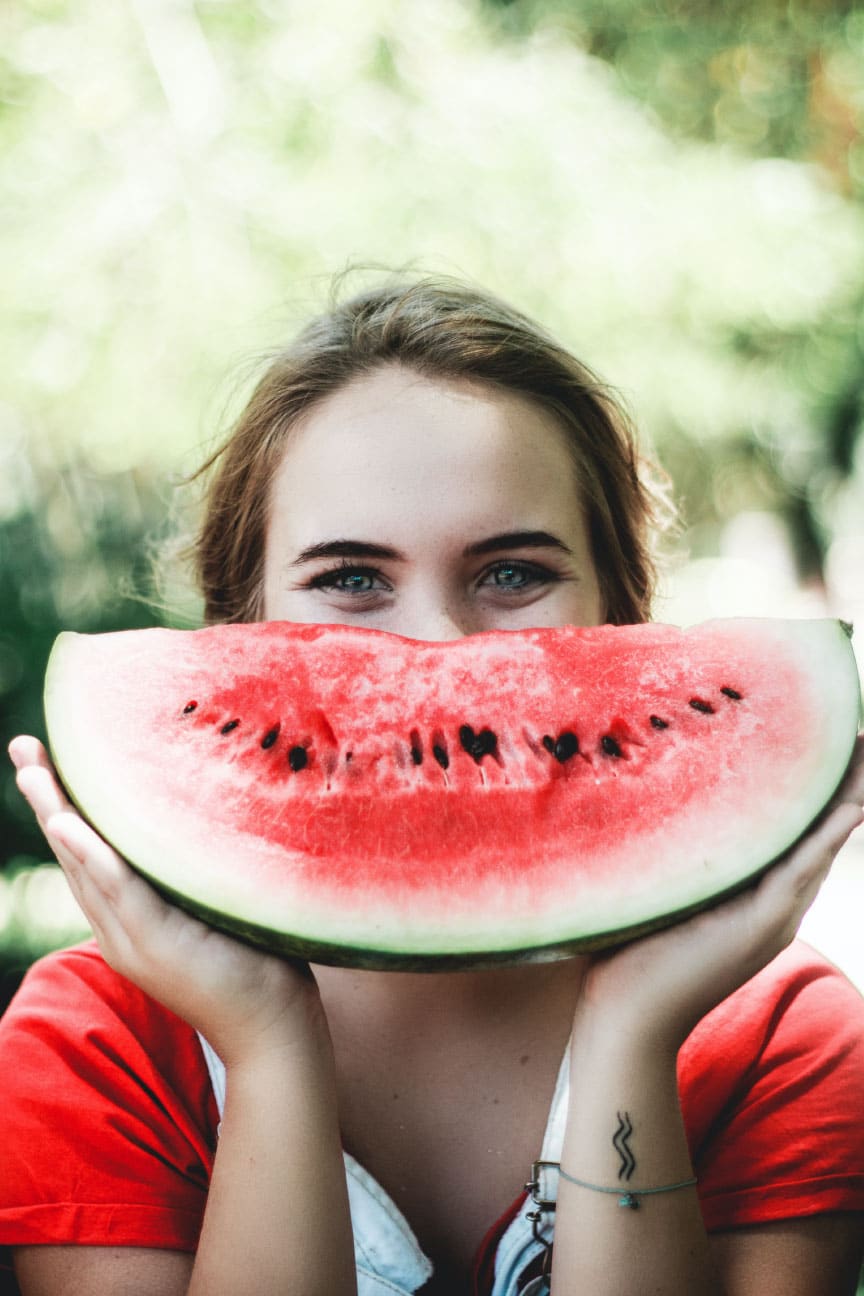 Woman holding a large slice of watermelon in front of her face, smiling, with a blurred green background.