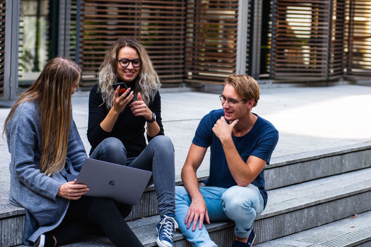 Group of three young adults discussing home loan modifications, one using a laptop, another holding a smartphone, in a modern outdoor setting.
