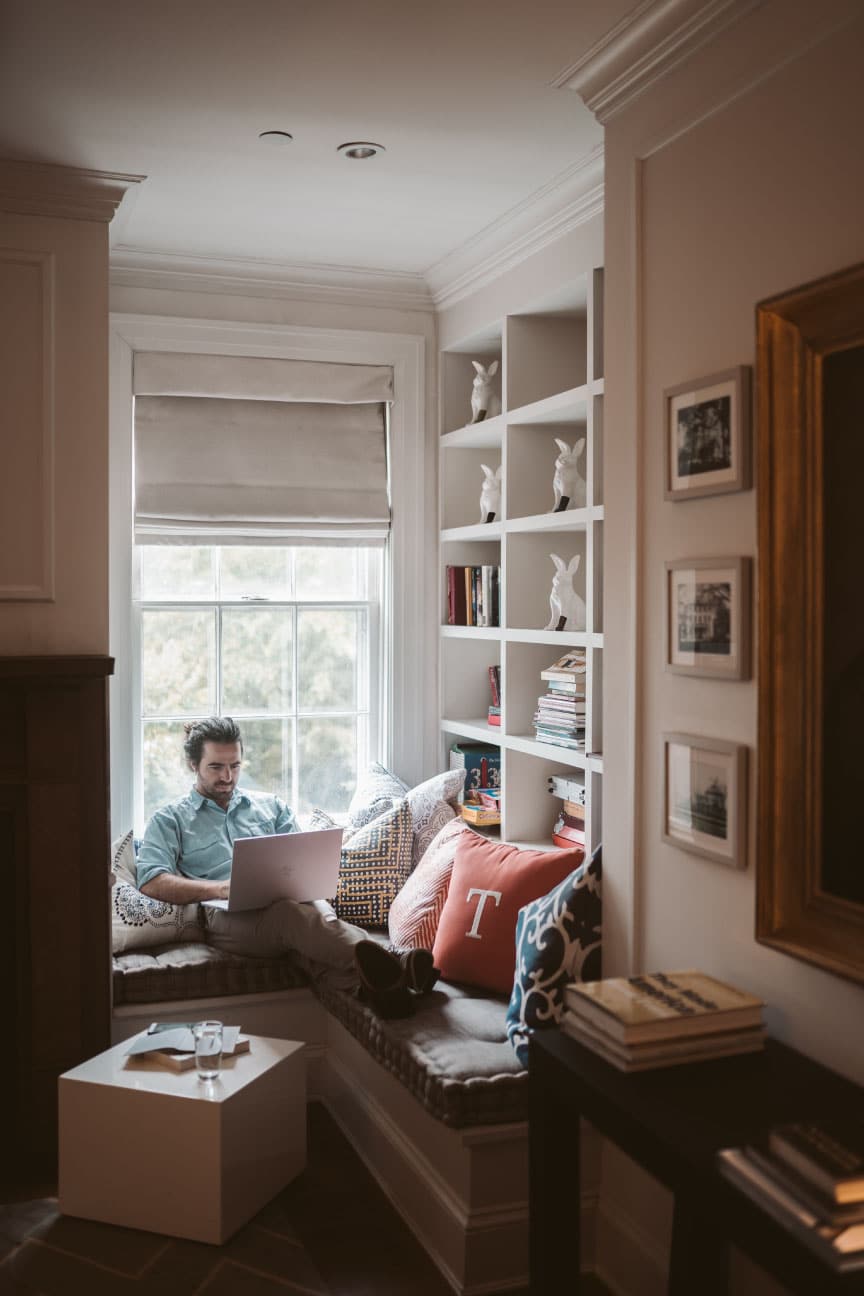 Man using a laptop in a cozy reading nook with decorative pillows and bookshelves, emphasizing home life and comfort for potential home buyers.