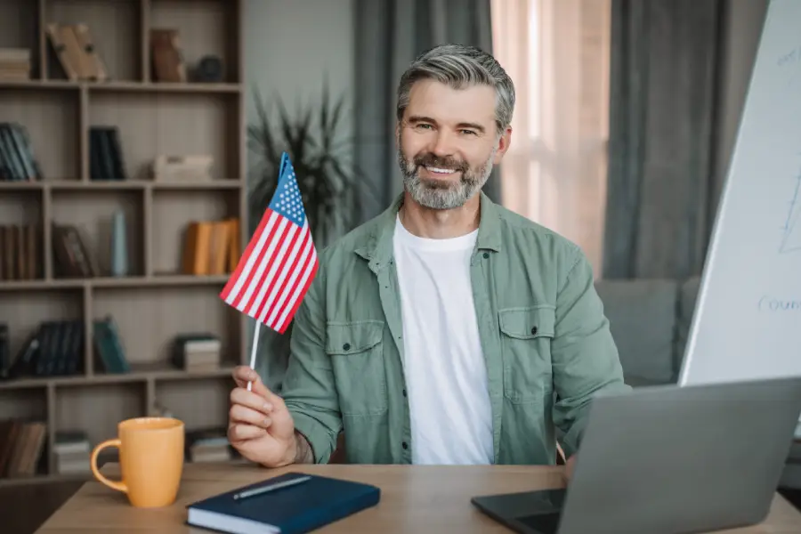 Smiling man holding a small American flag, sitting at a desk with a laptop, coffee mug, and notebook, representing support for VA home loans for veterans and military families.