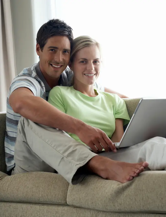 Happy couple sitting on a couch using a laptop, representing accessibility and support in exploring FHA home loan options.